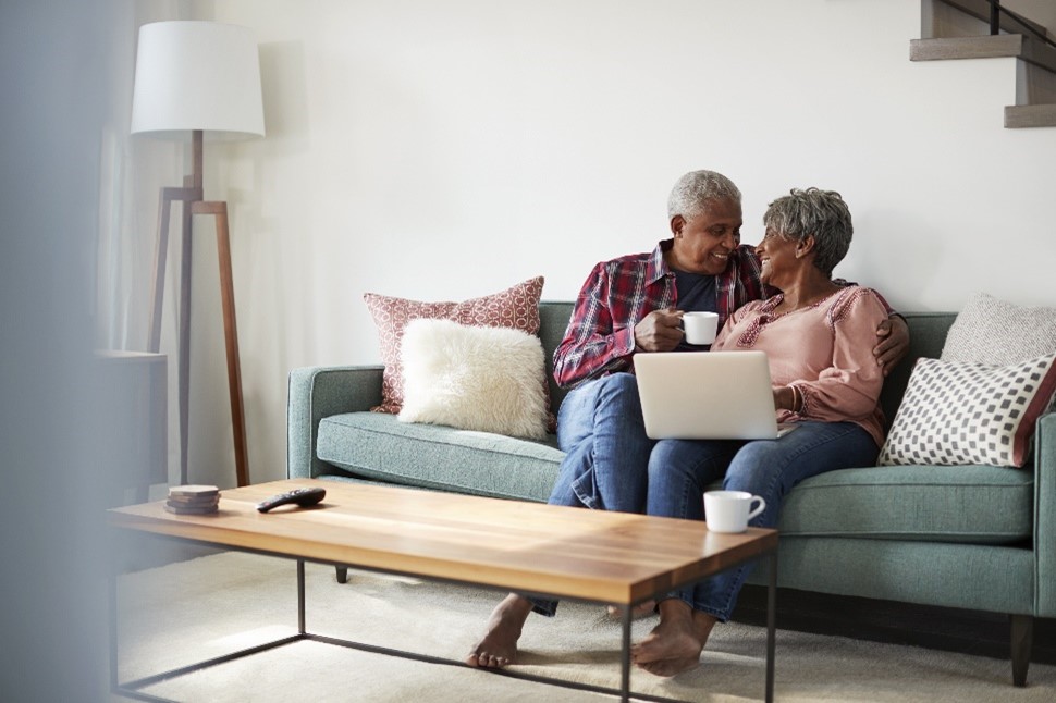 Image of an elderly husband and wife snugly sitting on a couch, looking over renovation plans on a laptop.