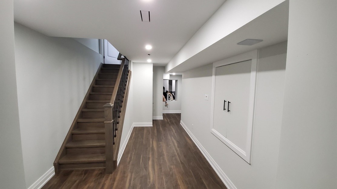 Image of a newly refinished basement with light-grey walls, white trim, and wood-look flooring.